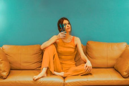 Affiliate Suitability - Full body of focused barefoot female wearing casual clothes sitting with bent leg and pot with flower near face on sofa near green wall in room